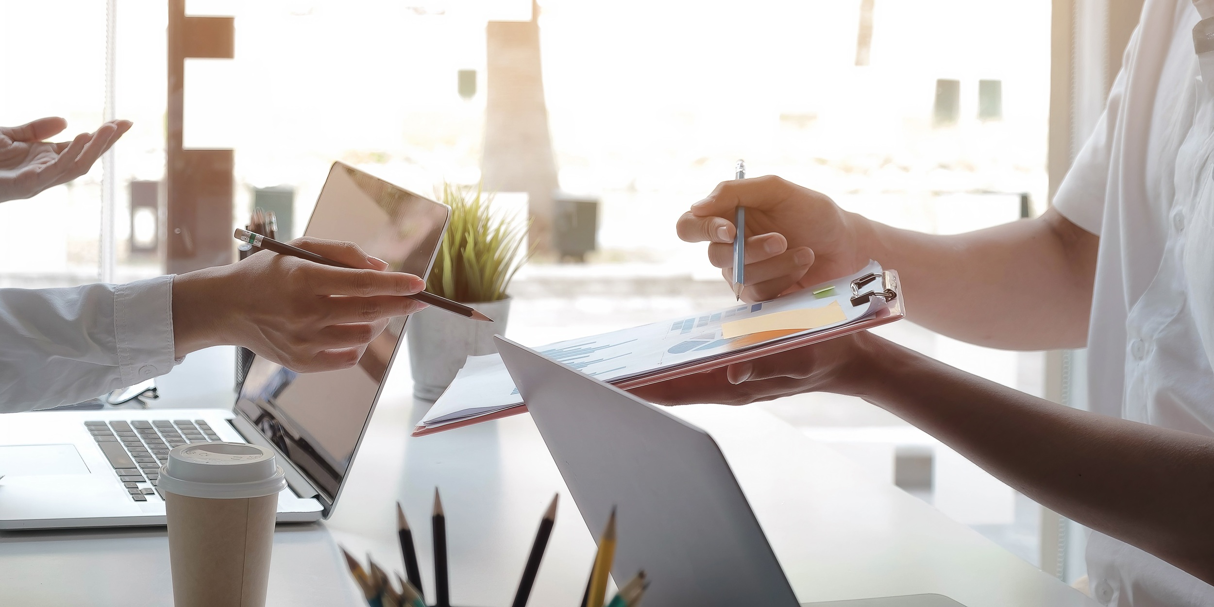 Woman points a pencil at a clipboard she is holding. On the clipboard are bar graphs and pie charts. A man's hand is seen pointing at the clipboard. There is a laptop in front of each person and az cup of coffee on the desk. Credit: Shutterstock