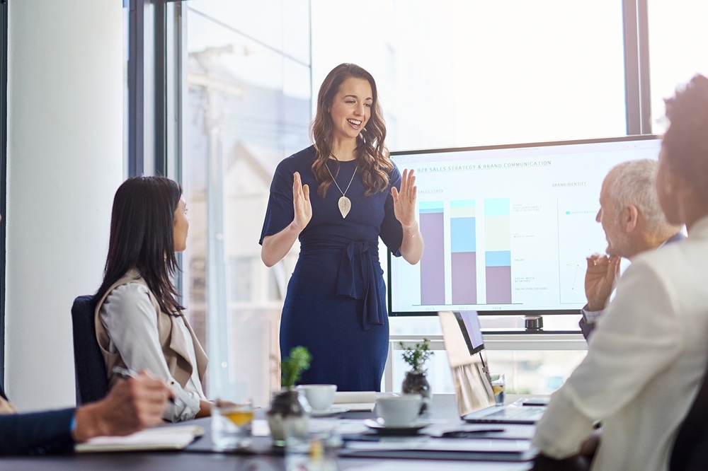 Businesswoman presents info to 4 people who are seated at a conference table. Credit: Shutterstock
