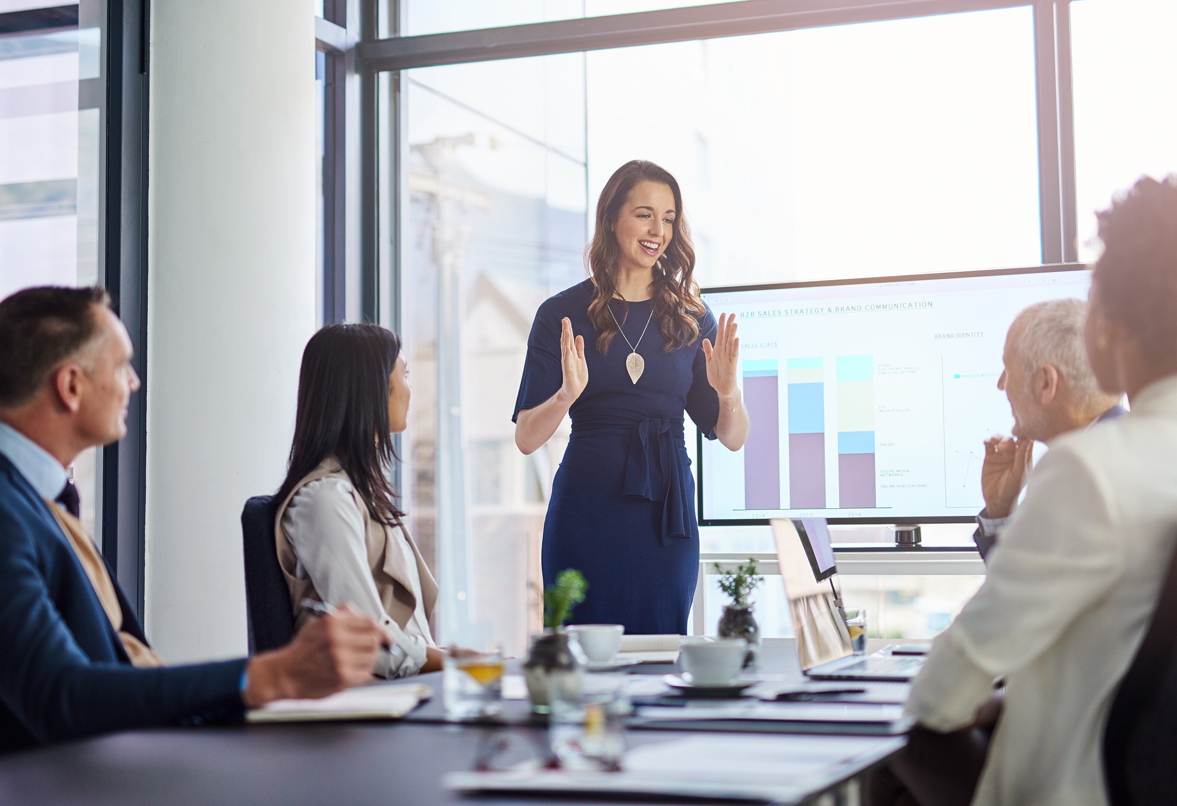 Businesswoman presents info to 4 people who are seated at a conference table. Credit: Shutterstock
