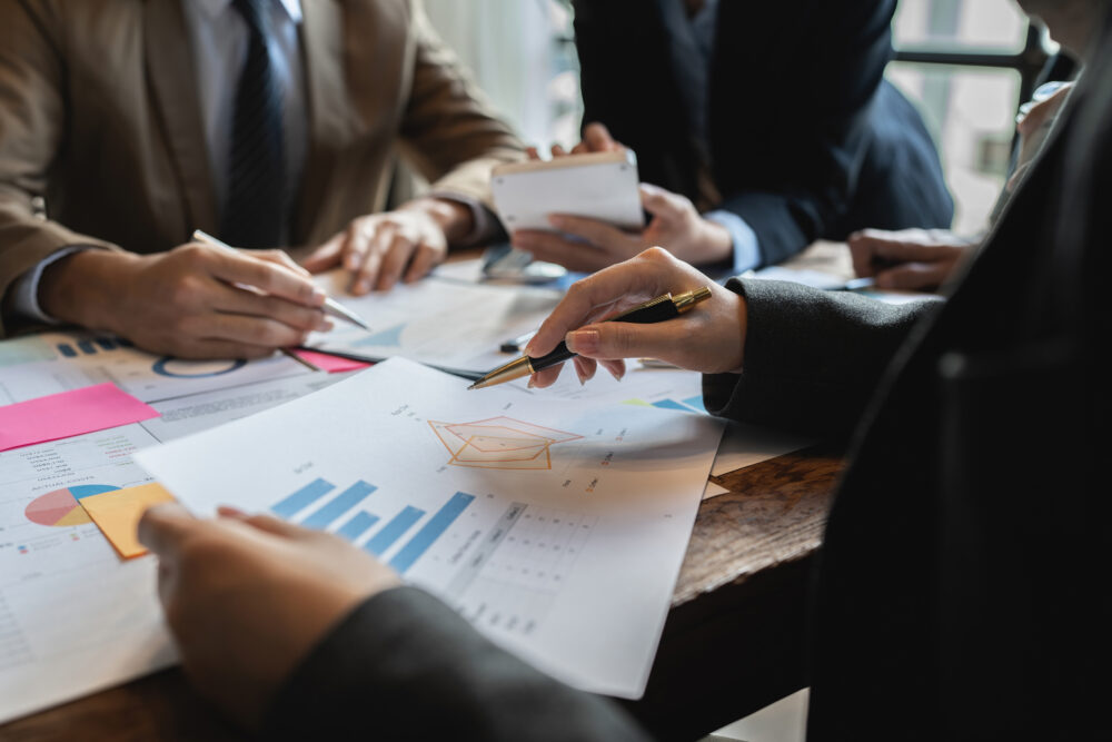 Three people at a table are holding documents with bar charts and graphs, point to the information with pens. Credit: iStock Photo.